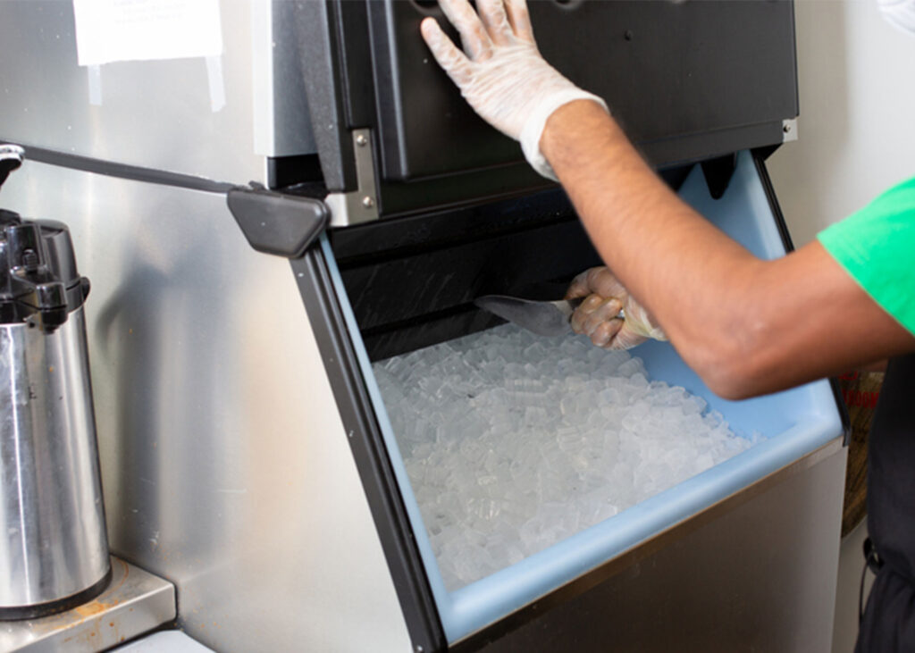 Person scooping ice from a commercial ice machine in a restaurant or kitchen setting.